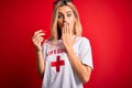 Young beautiful blonde lifeguard woman wearing t-shirt with red cross using whistle cover mouth with hand shocked with shame for