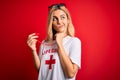 Young beautiful blonde lifeguard woman wearing t-shirt with red cross using whistle serious face thinking about question, very