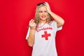 Young beautiful blonde lifeguard woman wearing t-shirt with red cross using whistle stressed and frustrated with hand on head,