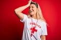 Young beautiful blonde lifeguard woman wearing t-shirt with red cross and whistle smiling confident touching hair with hand up