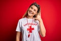 Young beautiful blonde lifeguard woman wearing t-shirt with red cross and whistle smiling doing phone gesture with hand and