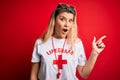 Young beautiful blonde lifeguard woman wearing t-shirt with red cross and whistle Surprised pointing with finger to the side, open