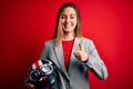 Young beautiful blonde motorcyclist woman holding motorcycle helmet over red background happy with big smile doing ok sign, thumb Royalty Free Stock Photo