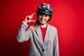 Young beautiful blonde motorcyclist woman wearing motorcycle helmet over red background smiling and confident gesturing with hand Royalty Free Stock Photo