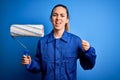 Young beautiful blonde painter woman with blue eyes painting wearing uniform using roller annoyed and frustrated shouting with Royalty Free Stock Photo