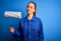 Young beautiful blonde painter woman with blue eyes painting wearing uniform using roller with a confident expression on smart Royalty Free Stock Photo