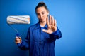 Young beautiful blonde painter woman with blue eyes painting wearing uniform using roller with open hand doing stop sign with Royalty Free Stock Photo