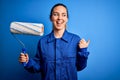 Young beautiful blonde painter woman with blue eyes painting wearing uniform using roller pointing and showing with thumb up to Royalty Free Stock Photo