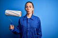 Young beautiful blonde painter woman with blue eyes painting wearing uniform using roller scared in shock with a surprise face, Royalty Free Stock Photo