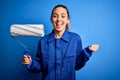 Young beautiful blonde painter woman with blue eyes painting wearing uniform using roller screaming proud and celebrating victory Royalty Free Stock Photo