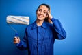 Young beautiful blonde painter woman with blue eyes painting wearing uniform using roller stressed with hand on head, shocked with Royalty Free Stock Photo