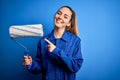 Young beautiful blonde painter woman with blue eyes painting wearing uniform using roller very happy pointing with hand and finger Royalty Free Stock Photo