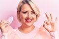 Young beautiful blonde plus size woman applying makeup using sponge over pink background doing ok sign with fingers, smiling Royalty Free Stock Photo