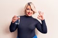 Young beautiful blonde plus size woman drinking glass of water over isolated white background doing ok sign with fingers, smiling Royalty Free Stock Photo