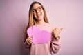 Young beautiful blonde romantic woman wearing glasses holding big pink heart pointing and showing with thumb up to the side with Royalty Free Stock Photo