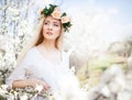 Young beautiful blonde smiling woman in white dress and rose wreath standing in blooming trees and looking aside Royalty Free Stock Photo