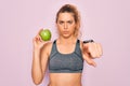 Young beautiful blonde sporty woman with blue eyes holding healthy green apple fruit pointing with finger to the camera and to Royalty Free Stock Photo