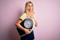 Young beautiful blonde sporty woman on diet holding weight machine over pink background cover mouth with hand shocked with shame