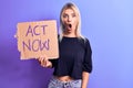 Young beautiful blonde woman asking for social reaction holding banner with act now message scared and amazed with open mouth for Royalty Free Stock Photo