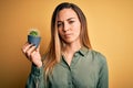 Young beautiful blonde woman with blue eyes holding small cactus pot over yellow bckground with a confident expression on smart Royalty Free Stock Photo