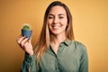 Young beautiful blonde woman with blue eyes holding small cactus pot over yellow bckground with a happy face standing and smiling Royalty Free Stock Photo