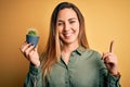 Young beautiful blonde woman with blue eyes holding small cactus pot over yellow bckground surprised with an idea or question Royalty Free Stock Photo