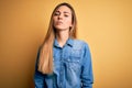 Young beautiful blonde woman with blue eyes wearing denim shirt over yellow background with serious expression on face Royalty Free Stock Photo