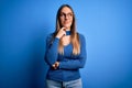 Young beautiful blonde woman with blue eyes wearing glasses standing over blue background with hand on chin thinking about Royalty Free Stock Photo