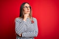 Young beautiful blonde woman with blue eyes wearing glasses standing over red background with hand on chin thinking about Royalty Free Stock Photo