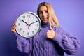 Young beautiful blonde woman doing countdown holding big clock over purple background happy with big smile doing ok sign, thumb up Royalty Free Stock Photo