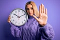 Young beautiful blonde woman doing countdown holding big clock over purple background with open hand doing stop sign with serious Royalty Free Stock Photo