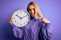 Young beautiful blonde woman doing countdown holding big clock over purple background with surprise face pointing finger to Royalty Free Stock Photo