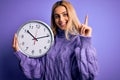 Young beautiful blonde woman doing countdown holding big clock over purple background surprised with an idea or question pointing Royalty Free Stock Photo