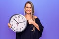 Young beautiful blonde woman doing countdown using big clock over purple background smiling happy and positive, thumb up doing Royalty Free Stock Photo