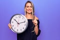 Young beautiful blonde woman doing countdown using big clock over purple background smiling with an idea or question pointing Royalty Free Stock Photo