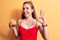 Young beautiful blonde woman drinking a glass of orange juice over yellow background doing ok sign with fingers, smiling friendly Royalty Free Stock Photo