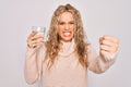 Young beautiful blonde woman drinking glass of water over isolated white background annoyed and frustrated shouting with anger, Royalty Free Stock Photo