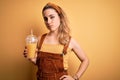 Young beautiful blonde woman drinking healthy glass of orange juice over yellow background with a confident expression on smart Royalty Free Stock Photo