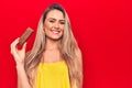 Young beautiful blonde woman eating chocolate protein bar over isolated red background looking positive and happy standing and Royalty Free Stock Photo