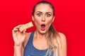 Young beautiful blonde woman eating muesli cereal bar over isolated red background scared and amazed with open mouth for surprise, Royalty Free Stock Photo