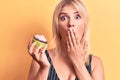 Young beautiful blonde woman eating sweet chocolate cupcake over isolated yellow background covering mouth with hand, shocked and Royalty Free Stock Photo