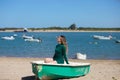 Young, beautiful blonde woman in an elegant green dress is sitting in a green fisherman\'s boat on the seashore. In the background Royalty Free Stock Photo