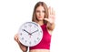 Young beautiful blonde woman holding big clock with open hand doing stop sign with serious and confident expression, defense Royalty Free Stock Photo