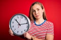 Young beautiful blonde woman holding big clock standing over isolated red background with a confident expression on smart face Royalty Free Stock Photo