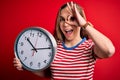 Young beautiful blonde woman holding big clock standing over isolated red background with happy face smiling doing ok sign with Royalty Free Stock Photo