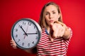 Young beautiful blonde woman holding big clock standing over isolated red background pointing with finger to the camera and to Royalty Free Stock Photo