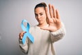 Young beautiful blonde woman holding blue cancer ribbon over isolated white background with open hand doing stop sign with serious Royalty Free Stock Photo