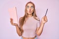 Young beautiful blonde woman holding book and pencil making fish face with mouth and squinting eyes, crazy and comical