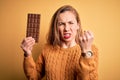 Young beautiful blonde woman holding chocolate bar standing over isolated yellow background annoyed and frustrated shouting with Royalty Free Stock Photo