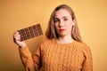 Young beautiful blonde woman holding chocolate bar standing over isolated yellow background with a confident expression on smart